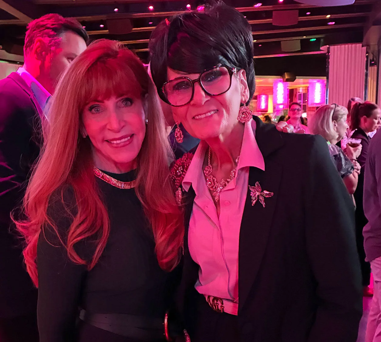 Two women stand close together and smile for the camera at an indoor social gathering. The setting features dramatic pink and red lighting with a crowd of people in the background. One woman wears a dark dress, while the other is dressed in a suit jacket,