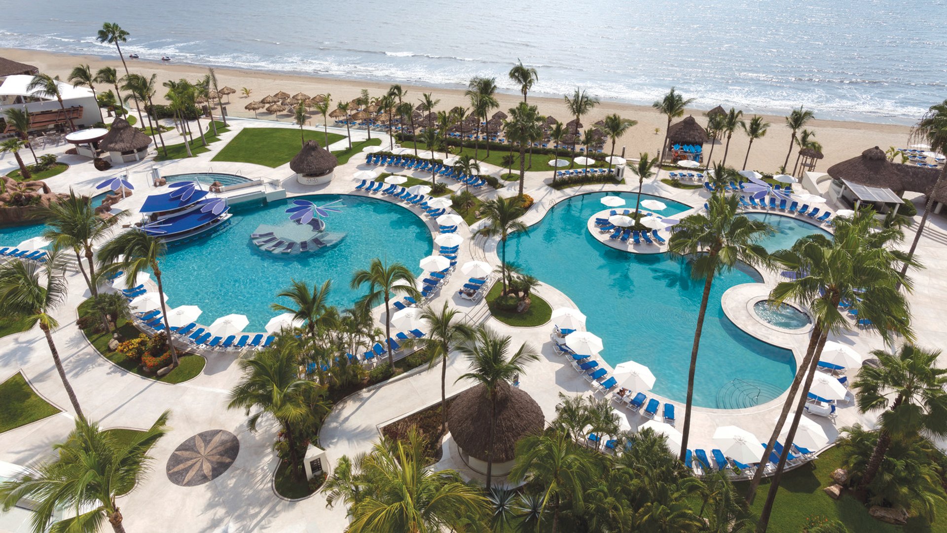 Aerial view of a resort with two large pools, lounge chairs, umbrellas, palm trees, and beach by the ocean.